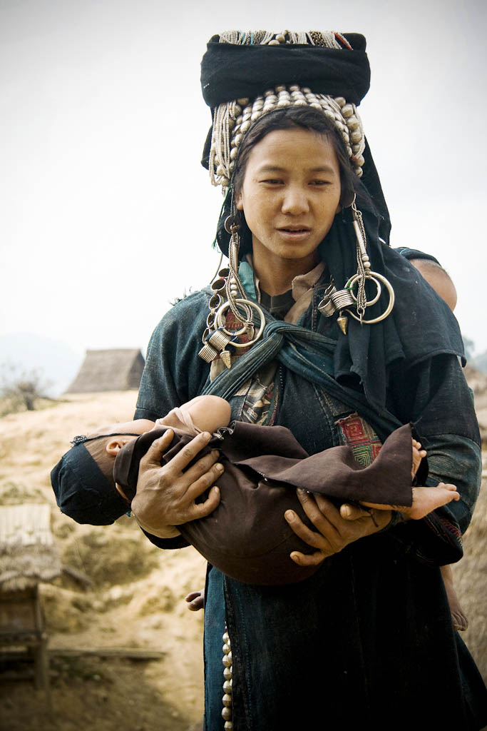 Young mother posing with her babies. The cotton fabric is not just used as shirt but also for instance to hold the baby in the back. She wears many different traditional Akha´s tribe complements made from silver. Only married women wear these silver ornaments, made with long time saved family money, which together with the traditional clothes get an unmistakable appearance. Akha people. Mountains Between Phongsali and Chinese border. Laos.
